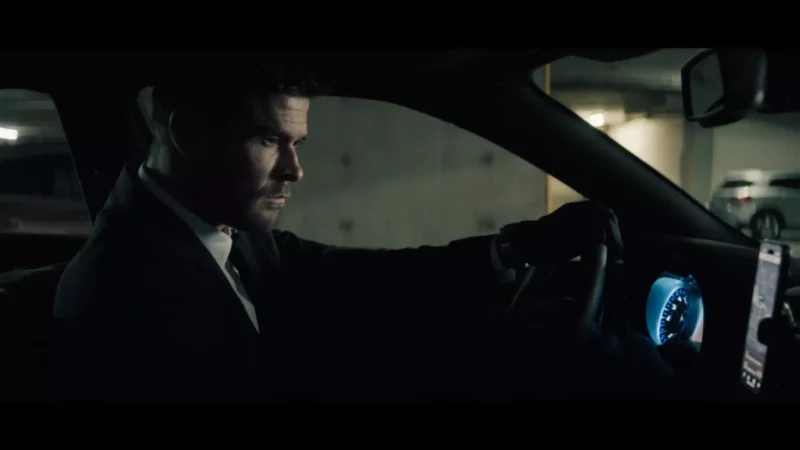 A man in a suit drives a car in a dimly lit parking garage. He looks focused and serious, gripping the steering wheel, with dashboard lights glowing.
