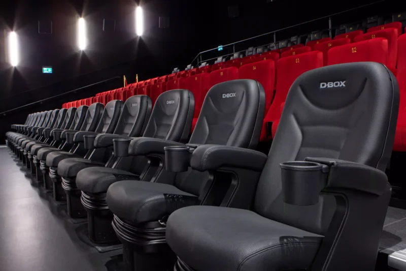 Rad čiernych pohyblivých sedadiel D-BOX v slabo osvetlenom kine s výraznými červenými sedadlami za nimi. Prostredie pôsobí moderne a pohlcujúco s dôrazom na pohodlie.