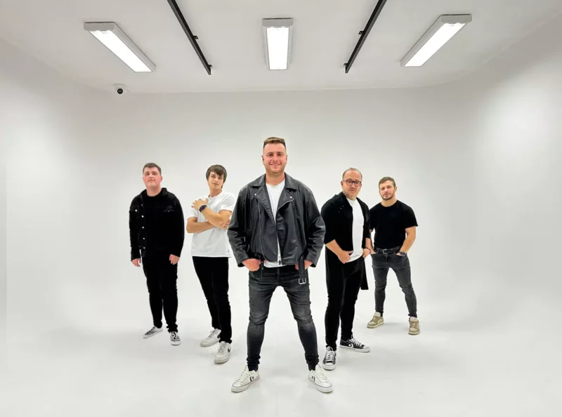 Päť mužov stojí v dobre osvetlenom bielom štúdiu. Ústredná postava má na sebe koženú bundu a vyžaruje sebavedomie. Skupina pôsobí ležérnou a jednotnou atmosférou.