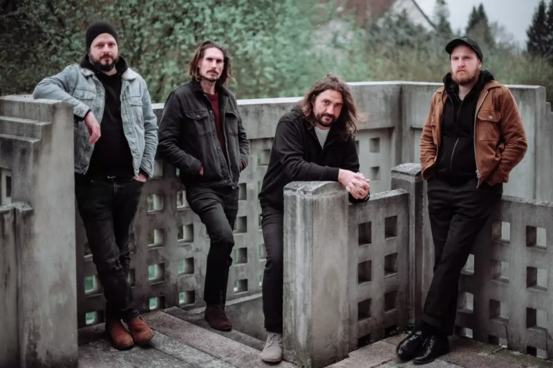 Four men stand on a concrete balcony, each leaning casually. The mood is relaxed, with trees in the background adding a natural setting.