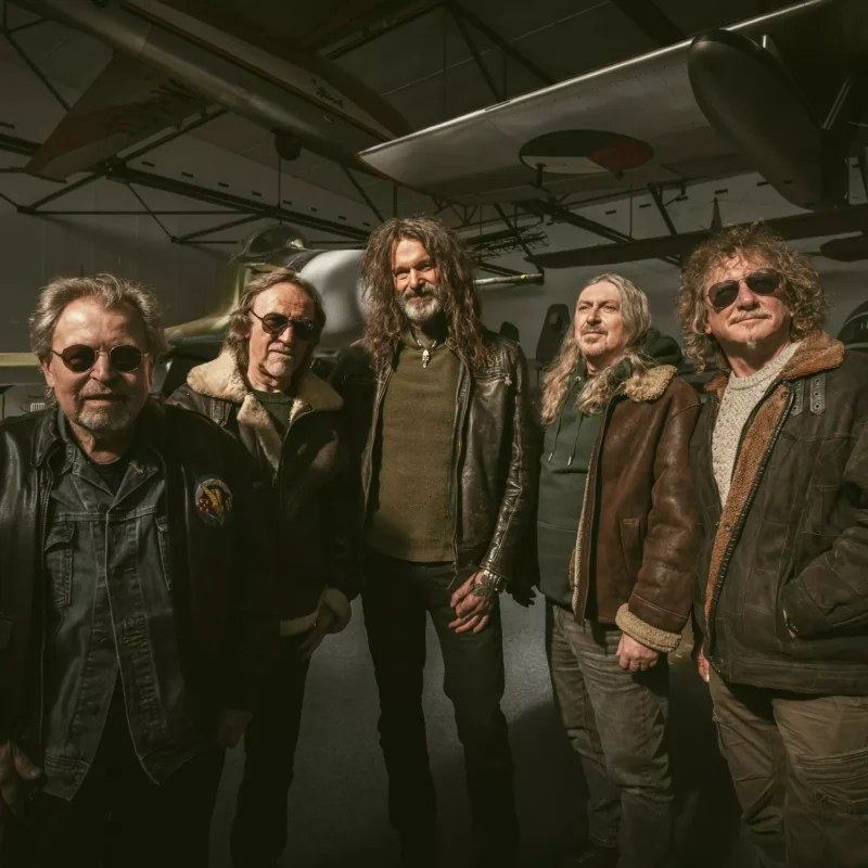 Päť mužov v kožených bundách sebavedomo stojí pod historickými lietadlami s vrtuľami a v slabo osvetlenom hangári vytvára dobrodružnú a nostalgickú atmosféru.