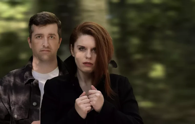 V lese s rozmazanou zeleňou stoja vážne vyzerajúci muž a žena. Žena je v popredí a zviera si tmavý kabát.