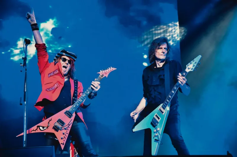 Dvaja gitaristi energicky hrajú na pódiu. Jeden má na sebe červenú bundu so zdvihnutou rukou, druhý v čiernej, obaja s farebnými gitarami na osvetlenom pozadí.