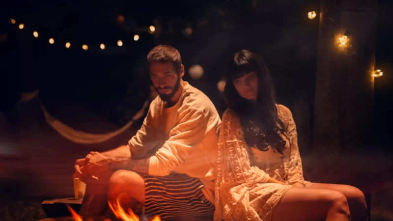 Pár sedí v noci chrbtom k sebe pri táboráku, obklopený svetelnými reťazami. Scéna je hrejivá a reflexívna, čo vytvára pokojnú a intímnu atmosféru.