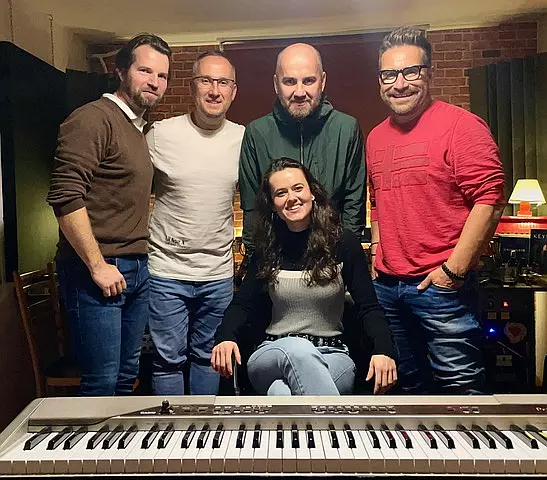 Five people in a music studio, four standing and one seated in front of a keyboard. They are smiling, conveying a warm, collaborative atmosphere.