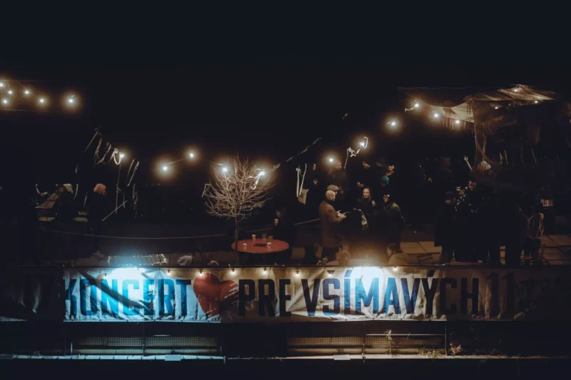 Nočná vonkajšia scéna s reťazovými svetlami osvetľujúcimi malú skupinu ľudí zhromaždených pri holom strome. Pod ňou je transparent s nápisom „KONCERT PRE VŠÍMAVÝCH“.
