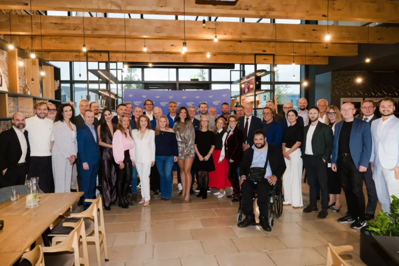 Veľká, rozmanitá skupina ľudí pózuje s úsmevom v modernej, príjemne osvetlenej miestnosti s drevenými trámami. Stoja spolu na formálnom stretnutí alebo oslave.