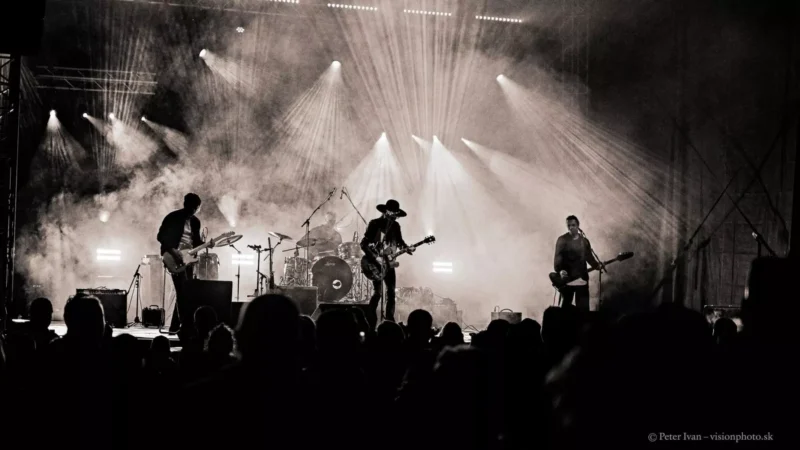 Silueta kapely energicky vystupuje na pódiu uprostred dynamického osvetlenia a dymu. Atmosféra je intenzívna a podmanivá, pričom sleduje scénu veľké publikum.