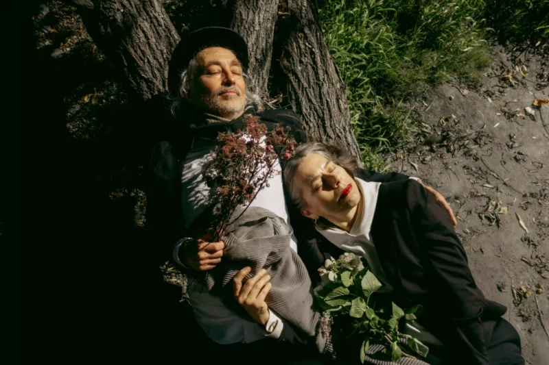 Starší pár odpočíva pod stromom so zatvorenými očami, každý drží kvety. Pôsobia pokojne, obklopení trávou a škvrnitým slnečným svetlom, čo vyžaruje pokoj.