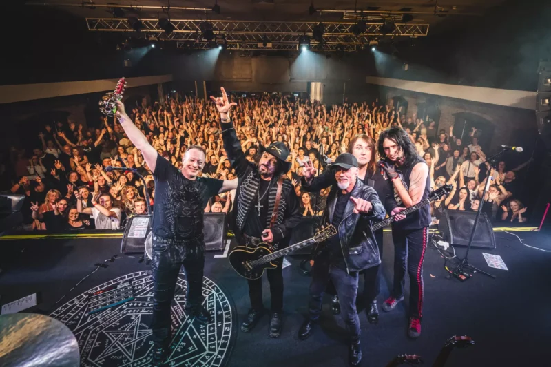 Rocková kapela na pódiu, usmievavá a pózujúca s gitarami, uprostred jásajúceho davu. Energická a triumfálna atmosféra s jasnými svetlami pódia a publikom dvíhajúcim ruky.