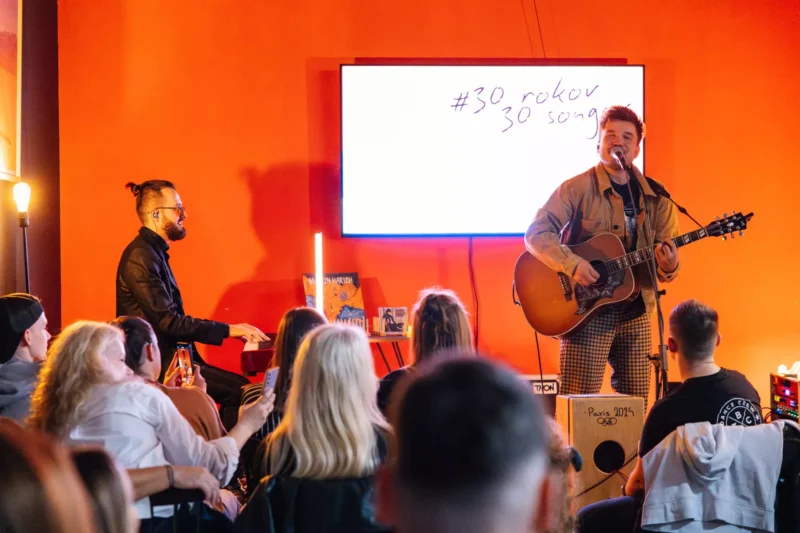 Gitarista spieva na pódiu, publikum sedí vnútri, pri oranžovej stene. Na obrazovke sa zobrazuje „#30 rokov 30 songov“. Útulná a intímna atmosféra.