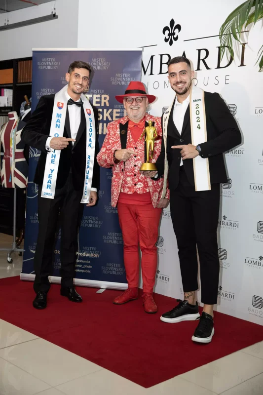 Traja muži stoja na červenom koberci. Osoba v strede má na sebe červený klobúk a kvetinový oblek, drží trofej, po bokoch sú dvaja muži v čiernych oblekoch so šerpami. Nálada je slávnostná.