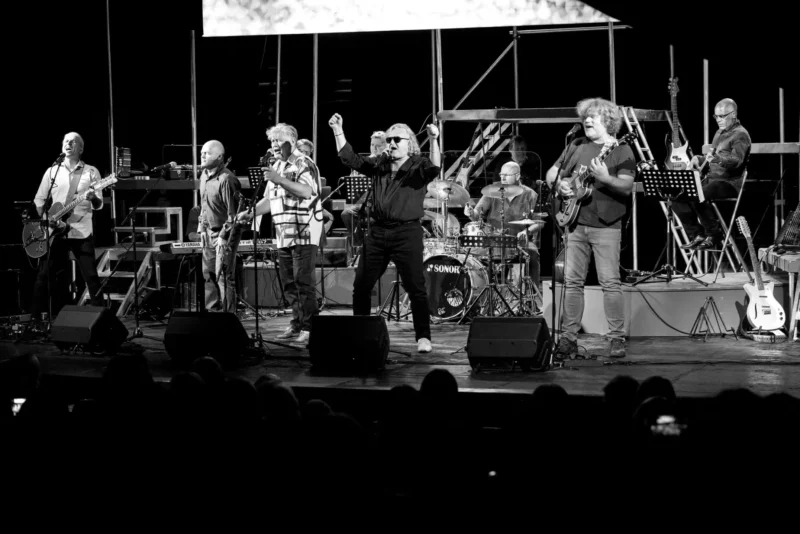Čiernobiely obraz živej kapely vystupujúcej na pódiu. Hudobníci hrajú na gitarách, bicích a vášnivo spievajú. Publikum je v tieni v popredí.