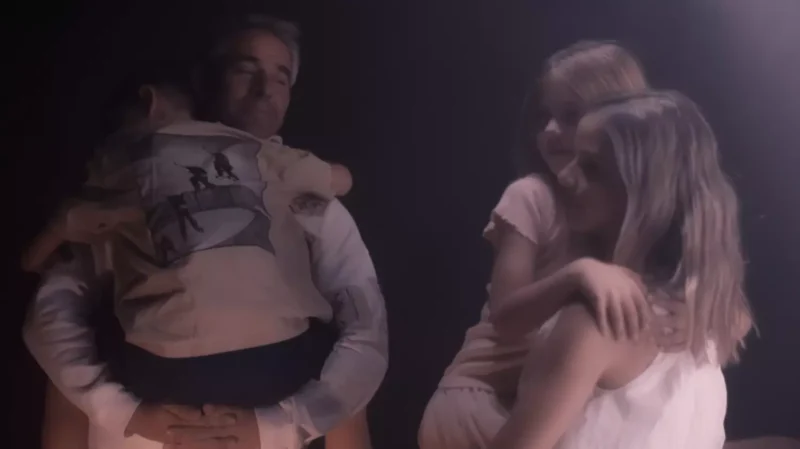 Rodina zažíva tlmené, nežné chvíle. Muž a žena držia malého chlapca a dievča. Nálada je pokojná a upokojujúca.