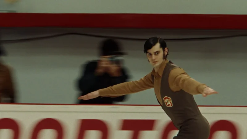 Krasokorčuliar v hnedom kostýme sa kĺže po klzisku s rozpaženými rukami. Fotograf v rozostrenom pozadí zachytáva tento okamih. Scéna pôsobí dynamicky a sústredene.