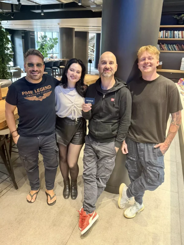 Štyria ľudia stoja blízko seba a usmievajú sa v modernej, jasne osvetlenej miestnosti. Vyzerajú uvoľnene a šťastne, ležérne oblečení, s poličkou na knihy a rastlinami v pozadí.