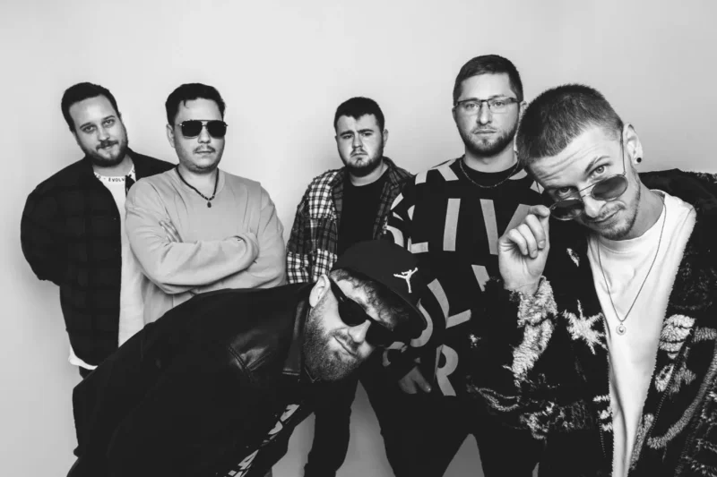 Čiernobiely obrázok šiestich mužov sebavedomo pózujúcich. Jeden muž sa nakláňa dopredu so slnečnými okuliarmi a vytvára tak neformálnu, chladnú atmosféru na jednoduchom pozadí.