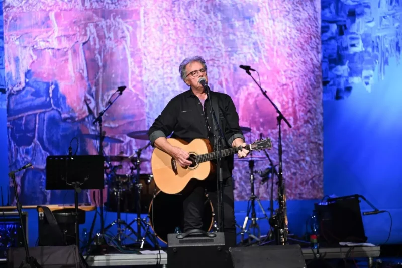 Hudobník v čiernom oblečení hrá na akustickej gitare a spieva na pódiu. Žiarivé pozadie je zmesou ružovej a modrej s pokojnou atmosférou.