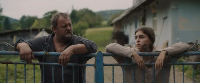 Muž a žena sa opierajú o modrú kovovú bránu a zamyslene hľadia do diaľky. Vidiecke prostredie zahŕňa dom a zelené polia pod zamračenou oblohou.