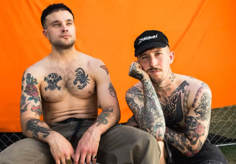 Dvaja potetovaní ľudia bez trička sedia na oranžovom pozadí. Jeden má na sebe čiernu čiapku a hlavu si opiera o ruku. Nálada je uvoľnená.