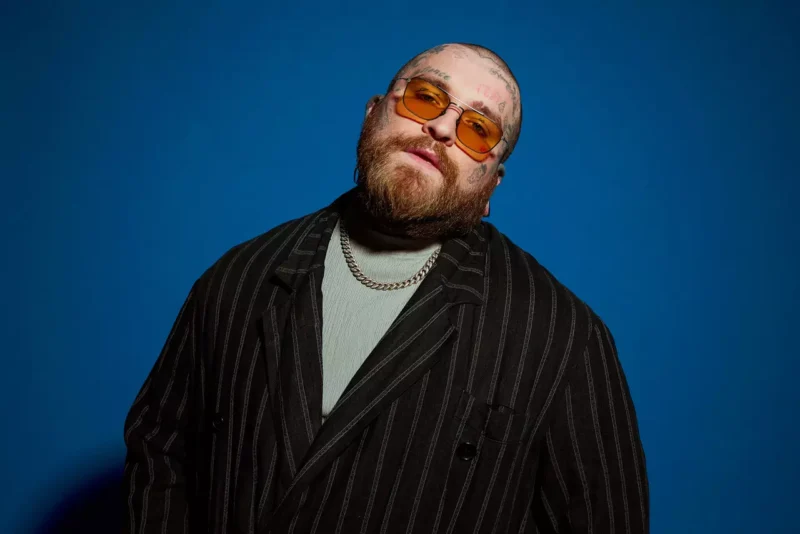 Bradatý muž s tetovaniami, v oranžových slnečných okuliaroch, pruhovanom obleku a so striebornou retiazkou, sebavedomo pózuje na modrom pozadí.