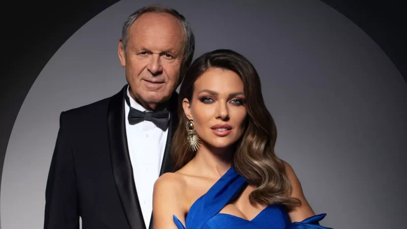 Starší muž v smokingu a žena v modrých šatách s prepracovanými náušnicami sebavedomo pózujú na tmavom, elegantnom pozadí.