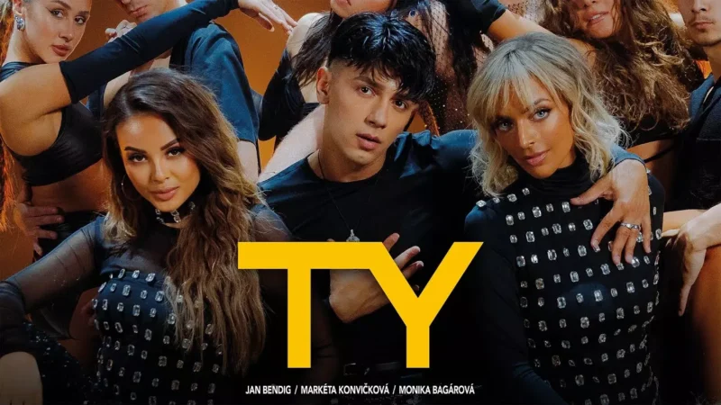 Skupina umelcov v čiernych outfitoch s cvočkami pózuje sebavedomo, obklopená tanečníkmi s dynamickými gestami. Navrchu je výrazný žltý nápis „TY“. Energická atmosféra.