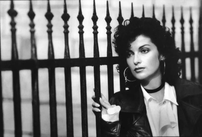 Čiernobiela fotografia ženy s kučeravými vlasmi a kruhovými náušnicami, ktorá zamyslene hľadí. Stojí pri kovanom železnom plote, má na sebe koženú bundu a košeľu s otvoreným golierom.