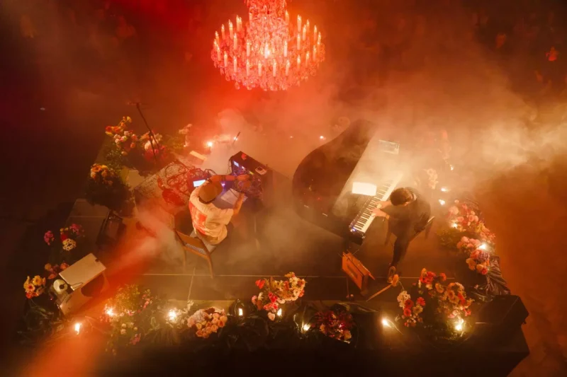 Aerial view of a dimly lit stage with two musicians, a piano, and electronic equipment. A grand chandelier casts a warm glow amid fog, surrounded by colorful flowers.
