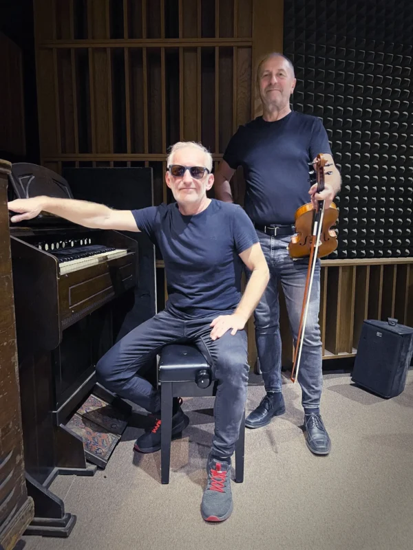 Dvaja muži v nahrávacom štúdiu; jeden sedí pri organe so slnečnými okuliarmi, zatiaľ čo druhý stojí a drží husle. Obaja majú na sebe čierne oblečenie a sebavedomo sa usmievajú.