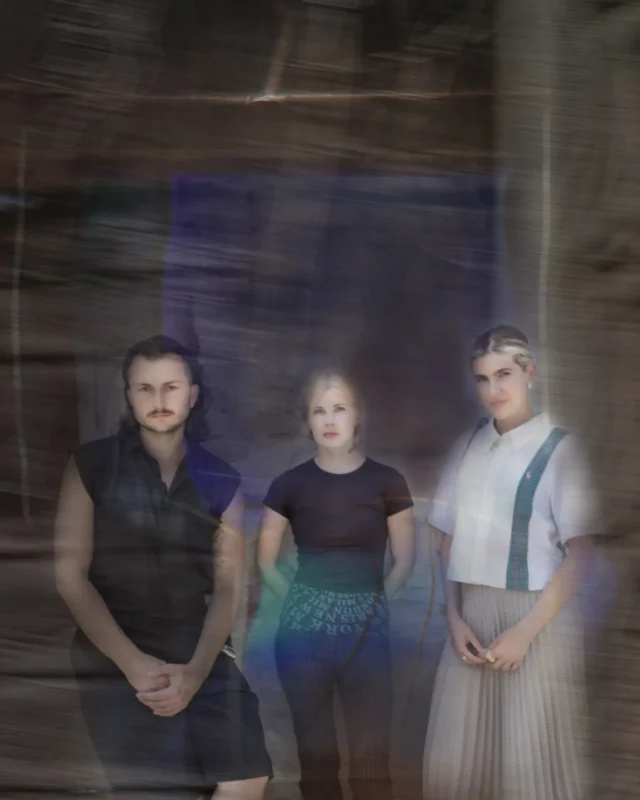 Traja jednotlivci stoja na rozmazanom, abstraktnom pozadí. Nálada je tajomná, s jemným osvetlením a éterickou atmosférou.