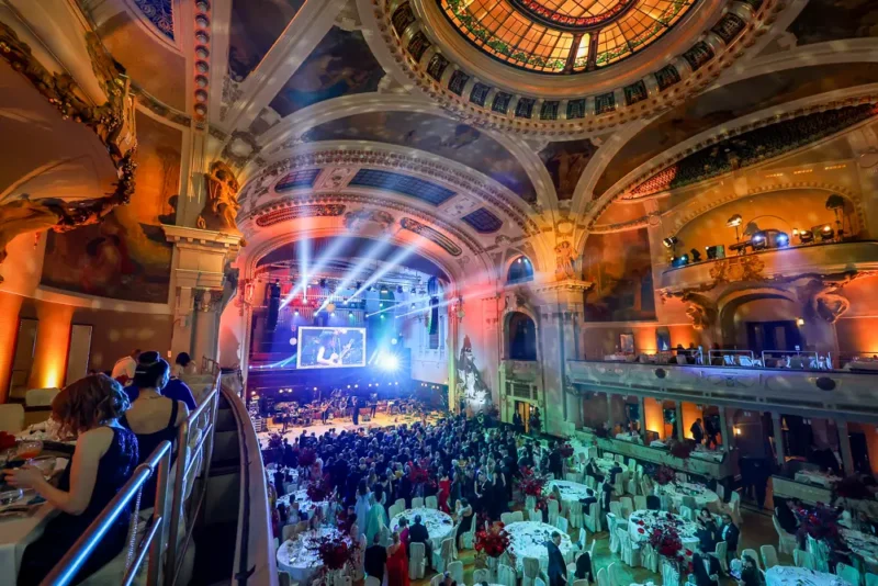 Elegantná tanečná sála so zdobenými stropmi, farebným osvetlením a živým davom. Formálna udalosť s okrúhlymi stolmi, výzdobou a živou hudbou na pódiu.