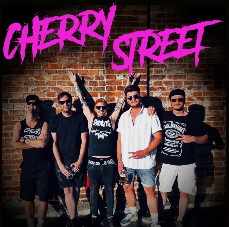 Päť mužov stojí pri tehlovej stene s výrazným ružovým graffiti „Cherry Street“ nad nimi. Sú ležérne oblečení a vyžarujú z nich zábavnú, rebelskú atmosféru.