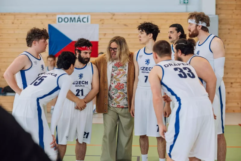 Basketbalový tím sa túli so svojím trénerom v telocvični a má na sebe biele dresy s nápisom „Česká republika“. Na drevenom pozadí visí česká vlajka.