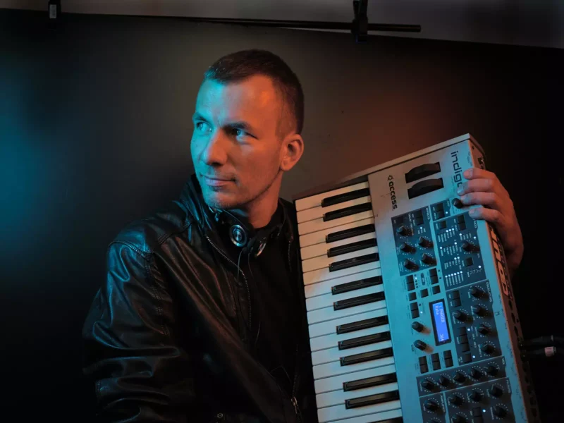 Muž v čiernej koženej bunde drží syntetizátor, okolo krku má slúchadlá. Zamyslene hľadí do strany na pozadí melancholického modrého pozadia.