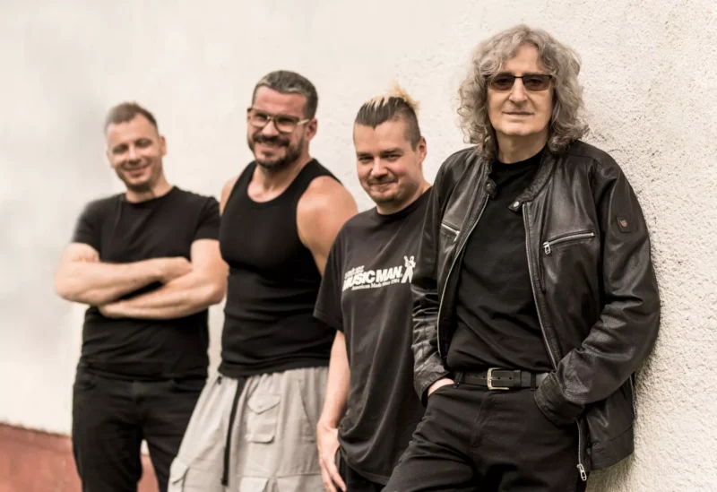 Štyria muži stoja ležérne pri svetlej stene. Traja majú na sebe čierne tričká, jeden v koženej bunde. Usmievajú sa a vyžarujú z nich uvoľnenú a priateľskú atmosféru.