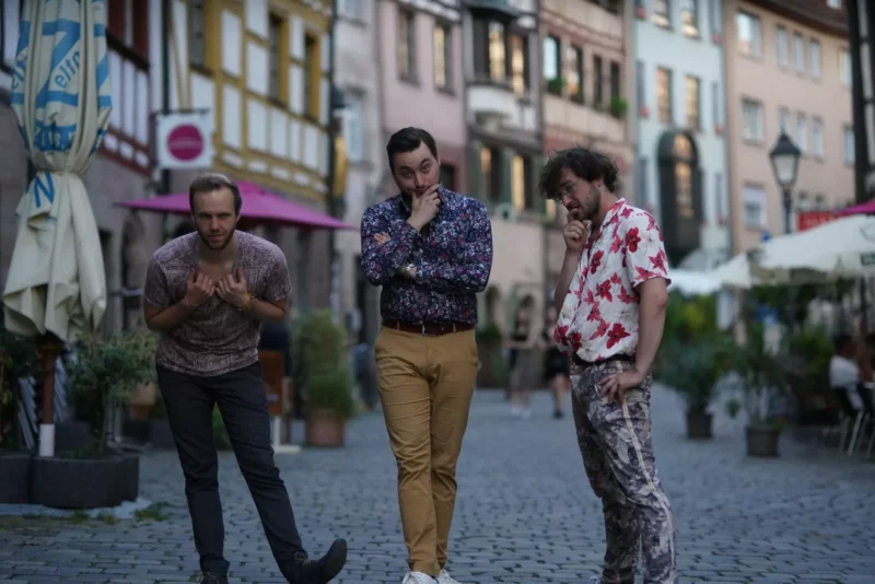 Traja muži hravo pózujú na dláždenej ulici lemovanej farebnými hrázdenými budovami. Nálada je bezstarostná a rozmarná.