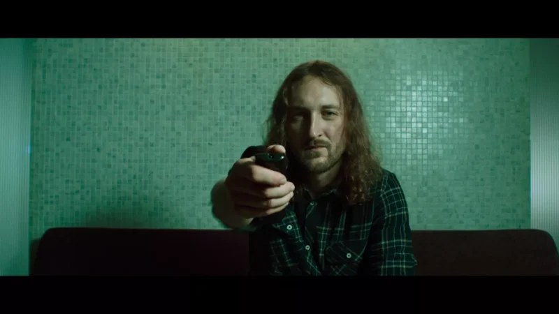 Muž s dlhými vlasmi a kockovanou košeľou sedí na gauči a drží diaľkové ovládanie nasmerované k divákovi. Pozadie tvorí svetlá mozaiková stena, ktorá vytvára uvoľnenú, kontemplatívnu atmosféru.