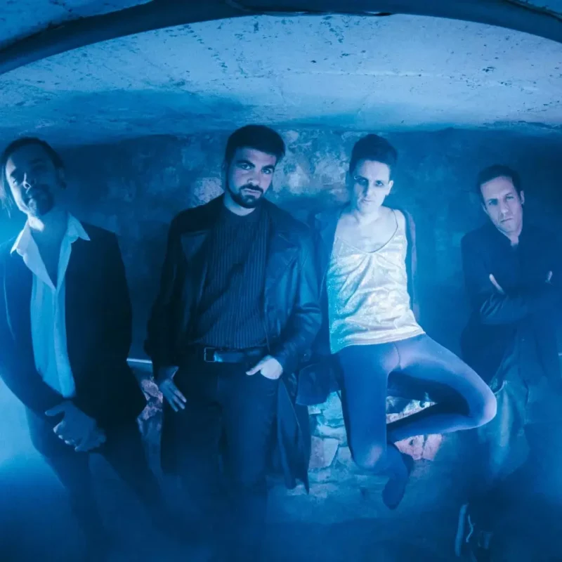 Štyria ľudia stoja v tmavej, modro osvetlenej miestnosti, ktorá vytvára tajomnú a melancholickú atmosféru. Jeden sedí, zatiaľ čo ostatní sebavedomo stoja okolo neho.