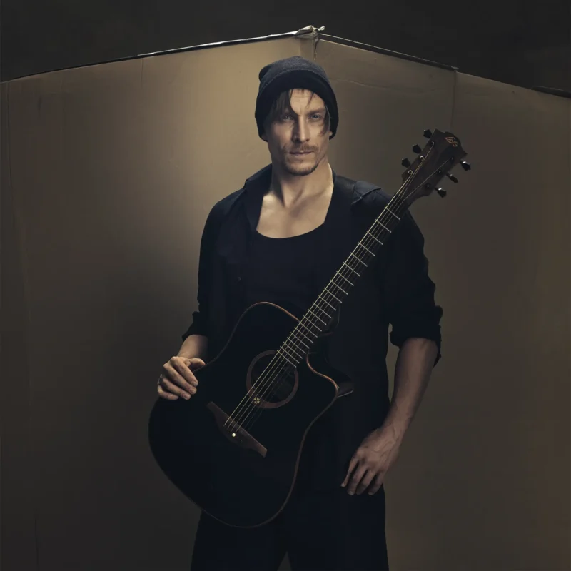 Muž v čiernej čiapke a bunde sebavedomo drží akustickú gitaru na jednoduchom pozadí, čím vytvára náladovú a umeleckú atmosféru.