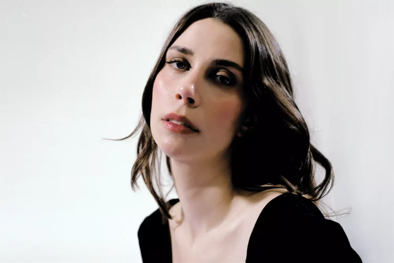 Žena s hnedými vlasmi po plecia jemne hľadí do kamery na čisto bielom pozadí. Má na sebe čierny top, ktorý vyjadruje pokojnú a introspektívnu náladu.