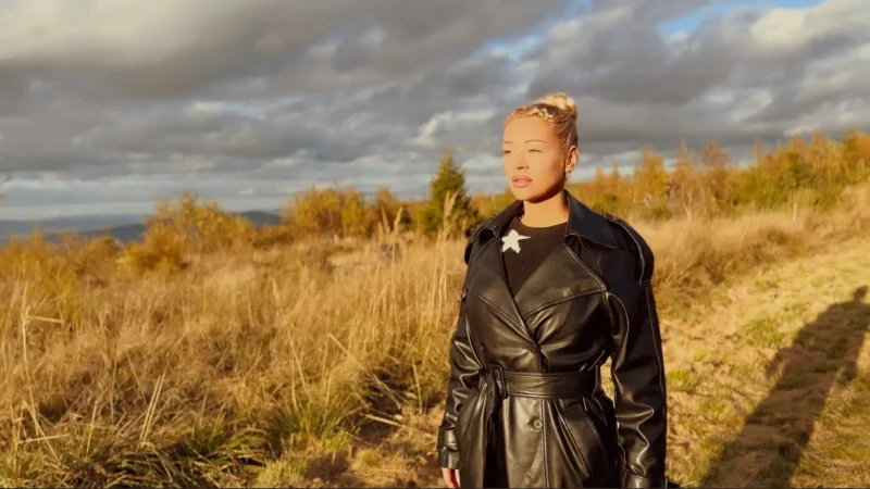 Žena v čiernom koženom kabáte stojí na slnkom zaliatom trávnatom poli pod dramatickou zamračenou oblohou a vyzerá zamyslene a pokojne.