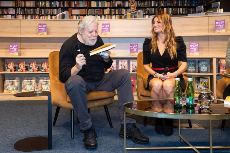 Starší muž s mikrofónom číta knihu vedľa usmievavej ženy v čiernych šatách. Sedia v útulnom kníhkupectve s vystavenými knihami.