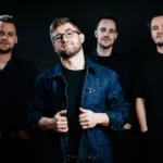 Štyria muži stoja na tmavom pozadí. Muž vpredu má okuliare a džínsovú bundu, ostatní majú na sebe čierne a vyžarujú z nich sebavedomie a kamarátstvo.
