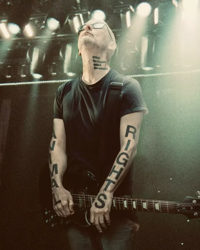 Človek stojí na pódiu pod jasnými svetlami a hrá na gitare. Na rukách má tetovania s nápisom „práva zvierat“. Pozerá sa hore s pokojným výrazom.