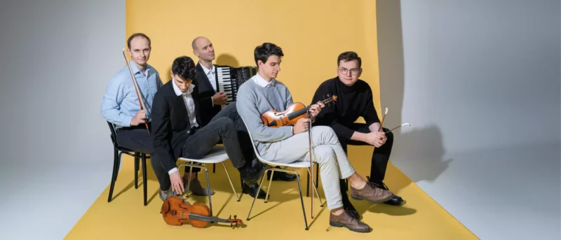 Päť mužov sedí na stoličkách na žltom pozadí. Držia hudobné nástroje, ako sú husle a akordeón, čím vytvárajú pokojnú umeleckú atmosféru.