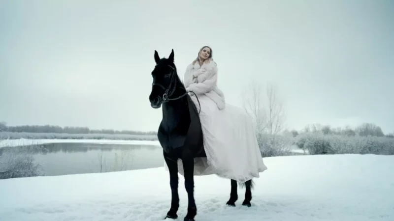 Žena v splývavých bielych šatách sedí na čiernom koni v zasneženej krajine pri pokojnom jazere a vyvoláva éterickú a pokojnú náladu.