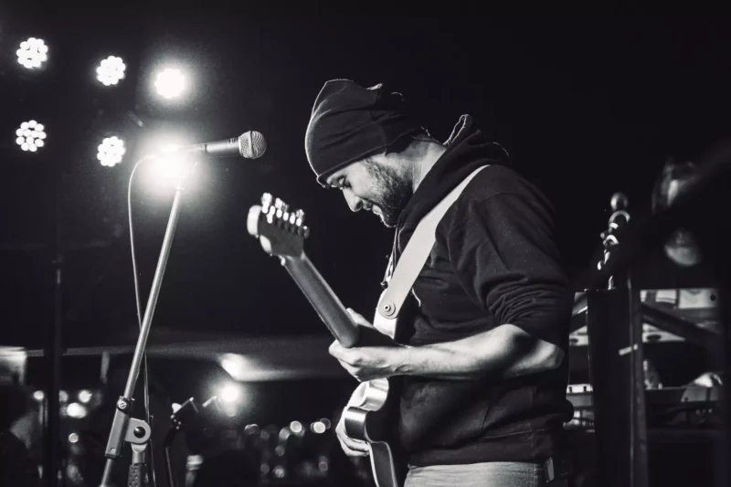 Black and white photo of a musician wearing a beanie, playing electric guitar on stage. Soft stage lights create a moody, intimate atmosphere.