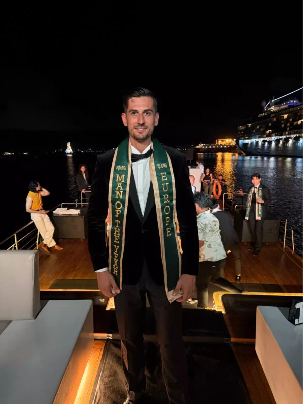 Muž v smokingu so šerpou s ocenením „Muž roka Európy“ stojí v noci na palube jachty. Živé pozadie s ľuďmi a svetlami mesta na vode.