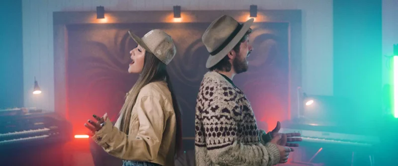 Dvaja ľudia stoja chrbtom k sebe v hudobnom štúdiu, obaja majú na sebe klobúky a sú oblečení v ležérnom oblečení. Zdá sa, že spievajú, obklopení hudobným vybavením pod farebným osvetlením.